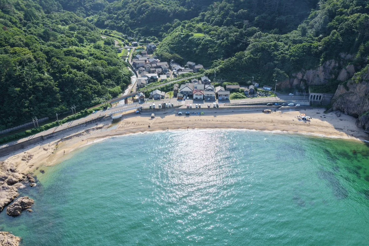 緑の山々に囲まれた入り江と砂浜、海沿いの集落を上空から見た風景